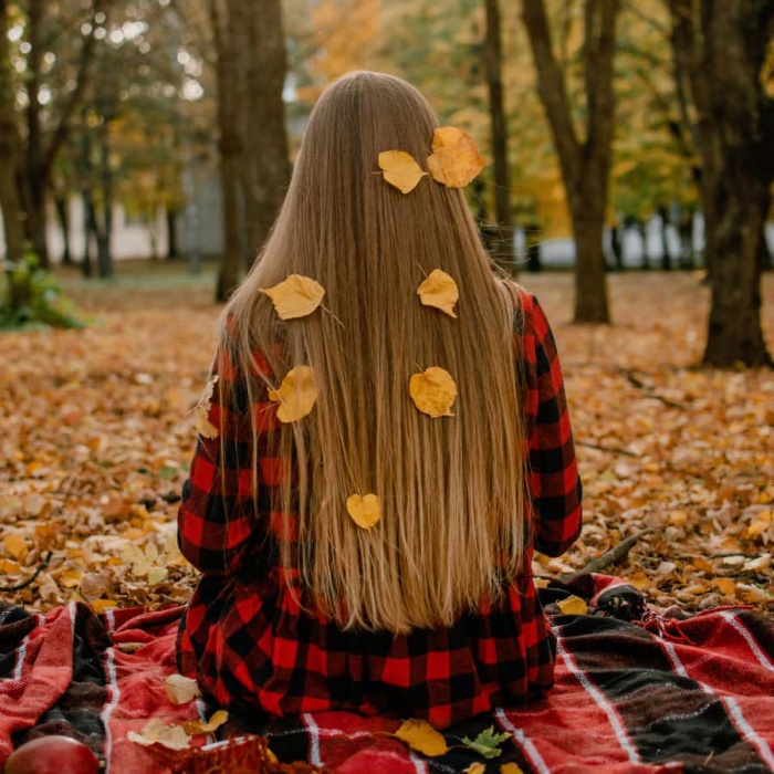 woman with long hair sitting on blanket as autumn leaves fall around her